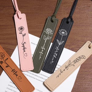 May include: Five leather bookmarks in various colors: brown, pink, green, black, and beige. Each bookmark features a floral design and a name in cursive script. The bookmarks have a matching leather strap.