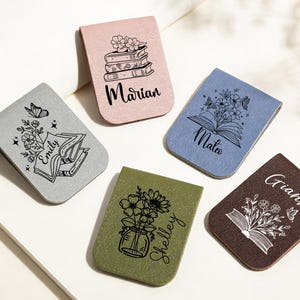 Puede incluir: Una colección de marcadores magnéticos personalizados en varios colores, incluyendo rosa, gris, azul, verde y marrón. Cada marcador presenta un diseño floral o temático de libros único con un nombre. Los marcadores están dispuestos sobre una superficie blanca.