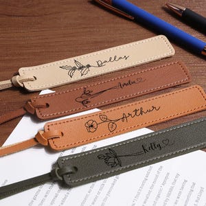 May include: Four personalised leather bookmarks in beige, brown, tan, and olive green, each with a name and floral design. The bookmarks have a stitched border and a matching leather tassel. Pens and a sheet of paper are in the background.