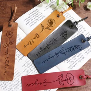 Pode incluir: Uma coleção de marcadores de couro personalizados em várias cores, incluindo castanho, bege, cinzento, azul e vermelho. Cada marcador apresenta um design floral e um nome, como "Arthur" ou "Joyce", com uma alça de couro.