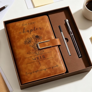 May include: A brown leather journal with the text "THE NEXT chapter" and "SOLEIL" with a floral design, a pen, and a ballpoint pen in a brown box. The journal has a gold-colored clasp. A cup of coffee and notepads are also visible.