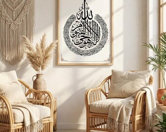 May include: A framed Islamic calligraphy art print in a light wood frame. The black calligraphy is centered on a white background. Two rattan chairs with cream-colored cushions and throws are in front of the print. A patterned rug and decorative wall hanging complete the scene.