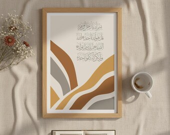 May include: A framed art print featuring Arabic calligraphy in a light beige frame. The artwork has a cream background with abstract wave-like shapes in shades of brown, gray, and gold. A cup and an open book are also visible.