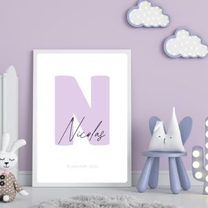May include: A framed print featuring a large lavender letter "N" with the name "Nicolas" in script. The date "10 January 2026" is below the name. The print is in a nursery setting with a crib, stuffed bunny, and decorative cloud and cactus lights.