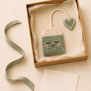 May include: A crocheted tea bag ornament in sage green and cream yarn, featuring a smiling face with closed eyes and pink cheeks. A cream-colored string connects the tea bag to a small, sage green heart-shaped tag, all presented in a brown box.