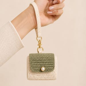 May include: A cream-colored crochet case for wireless earbuds featuring an olive green flap, a single white pearl-like button closure, and a matching cream-colored wrist strap attached with a gold-toned metal lobster clasp and ring.