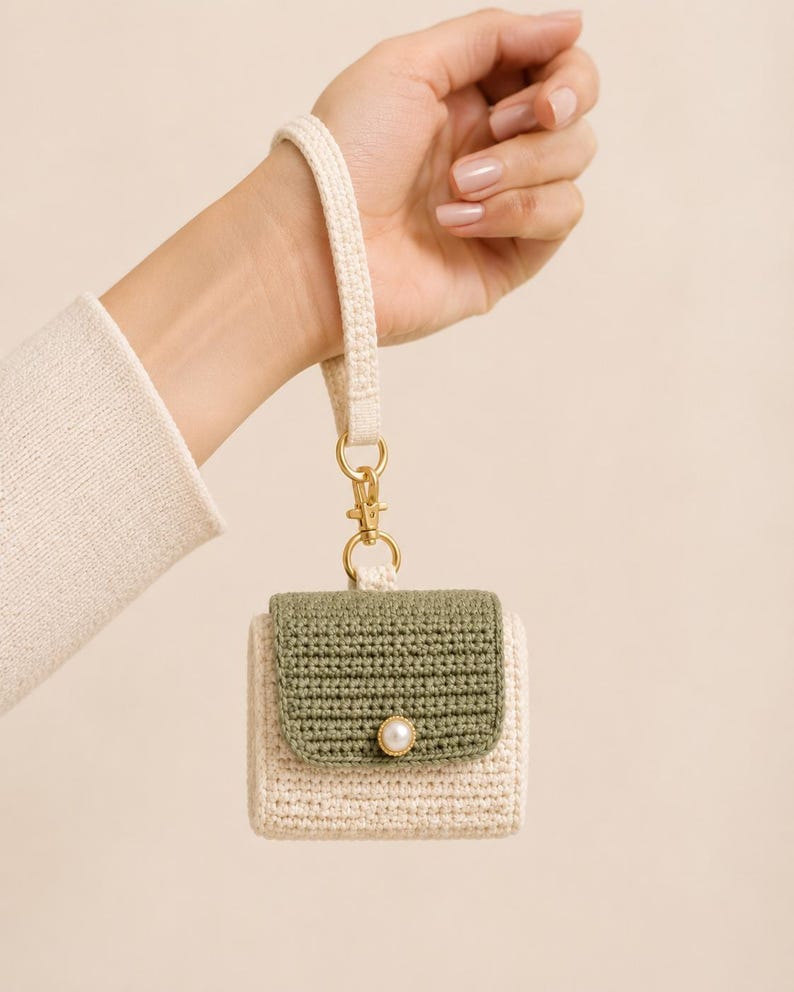 May include: A cream-colored crochet case for wireless earbuds featuring an olive green flap, a single white pearl-like button closure, and a matching cream-colored wrist strap attached with a gold-toned metal lobster clasp and ring.