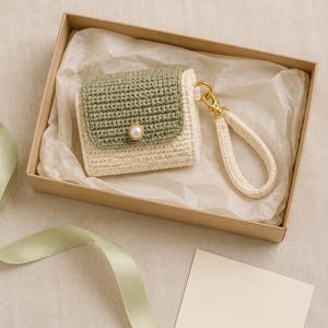 May include: A small, rectangular crochet pouch featuring a sage green flap and a cream-colored body. The flap is secured with a single white faux pearl button. A matching cream-colored crochet wrist strap is attached via a gold-toned metal clasp.