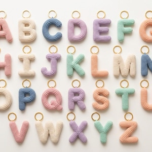 Puede incluir: Una colección de veintiséis llaveros individuales con letras del alfabeto, cada uno hecho de hilo de ganchillo en varios colores pastel como rosa, azul, crema, lavanda y verde menta, con una anilla de metal dorado sujeta en la parte superior.