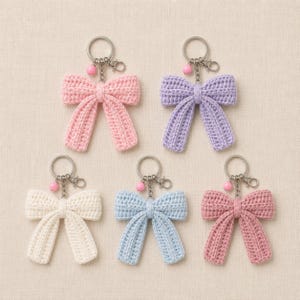 Può includere: Cinque portachiavi a forma di fiocco realizzati all'uncinetto disposti in gruppo. Ogni fiocco è fatto di filato in un colore solido diverso: rosa chiaro, lavanda, crema, azzurro e rosa antico. Ogni portachiavi ha un anello in metallo argentato e una piccola perlina rosa.