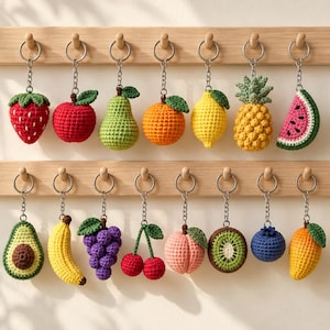 May include: A collection of fifteen crocheted fruit and vegetable keychains hanging from two wooden racks. The items include a strawberry, apple, pear, orange, lemon, pineapple, watermelon slice, avocado, banana, grapes, cherries, peach, kiwi, blueberry, and mango.