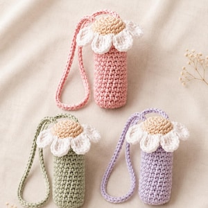 May include: Three crochet lipstick holders with daisy flower toppers. Each holder features a cylindrical body in pink, sage green, or lavender, topped with white petals and a beige center, and includes a matching crocheted wrist strap.