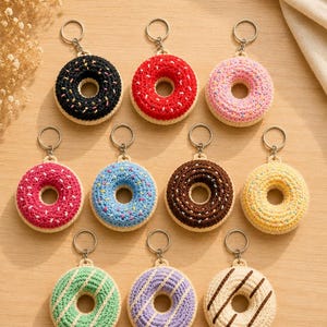 Könnte beinhalten: Eine Sammlung von elf gehäkelten Donut-Schlüsselanhängern in verschiedenen Farben wie Schwarz, Rot, Pink, Blau, Braun, Gelb, Grün, Lila und Creme, jeweils mit einem Metallring und gestickten Streuseln oder Glasurdetails versehen.