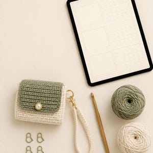 May include: A small crochet pouch with a sage green flap and cream body, featuring a pearl-like button, a matching cream wrist strap with a gold clasp, two balls of yarn, a wooden crochet hook, and four small green stitch markers.