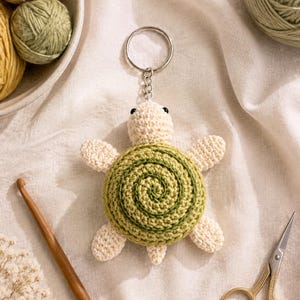 Peut inclure: Un porte-clés tortue au crochet avec une tête et des membres de couleur crème, doté d'une carapace ronde en spirale dans des tons de vert clair et foncé. La tortue est fixée à un anneau brisé en métal argenté par une courte chaîne en maillons.