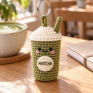 Puede incluir: Un llavero de ganchillo con forma de bebida matcha verde con una capa de nata montada blanca, una pajita pequeña y una cara sonriente. La parte delantera tiene un parche circular blanco con la palabra MATCHA bordada en hilo verde y una anilla.