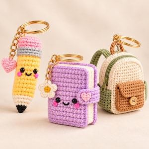 Può includere: Tre portachiavi all'uncinetto composti da una matita gialla con ciondolo a cuore rosa, un libro viola con ciondolo a fiore bianco e uno zaino beige e verde con tasca marrone e bottone in legno, fissati ad anelli in metallo color oro.