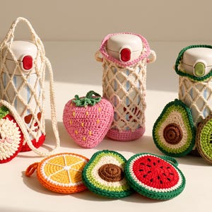 May include: Three white insulated water bottles encased in cream-colored crochet net carriers with straps, accompanied by several crocheted fruit-shaped coasters and pouches including an apple, strawberry, avocado, orange, and watermelon slice.