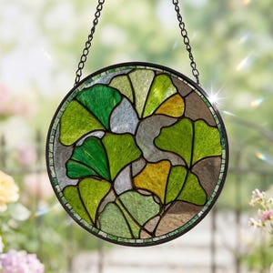 May include: Round stained glass window hanging with a black metal frame and chain. The design features green and yellow ginkgo leaf patterns. The window is displayed in front of a blurred background of flowers and greenery.