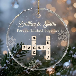 May include: A clear, round Christmas ornament with the text "Brothers & Sisters Forever Linked Together." The ornament features a Scrabble-style design with names spelled out. It hangs from a gold string, surrounded by festive decorations.