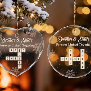 May include: Two clear glass Christmas ornaments, one heart-shaped and one round, hanging from a tree. Both ornaments have the text "Brothers & Sisters Forever Linked Together" and personalized names in a crossword style. Snowflakes are also featured.