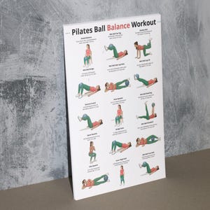 May include: A white Pilates Ball Balance Workout poster with illustrations of exercises. The poster features a variety of exercises, including seated balance, mini ball toe tap, and donkey kick. The poster is displayed against a gray wall.