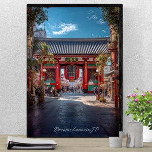 Op de afbeelding: Een ingelijste prent van een Japanse tempelpoort, met een grote rode lantaarn en traditionele architectuur. De afbeelding toont een drukke straatscène met mensen die onder een blauwe hemel naar de tempel lopen. De prent wordt weergegeven op een houten oppervlak.