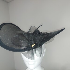 May include: A black formal hat with a wide brim and a decorative bow. The hat features a mesh overlay with small, sparkling accents. A cluster of gold-colored beads adorns the center of the bow. This elegant headwear is suitable for special occasions.