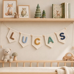 May include: A decorative banner featuring five off-white fabric pennants hanging from a string, each displaying a single knitted letter in brown, light blue, yellow, green, and dark blue, spelling the name LUCAS, accented with wooden beads.