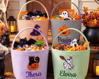 Może przedstawiać: Cztery materiałowe wiaderka na Halloween w pionowe paski w kolorach brzoskwiniowym, szarym, lawendowym i jasnym zielonym. Każde wiaderko ma wyhaftowane kursywą imię oraz ozdobną naszywkę z duchem, dynią lub kotem.