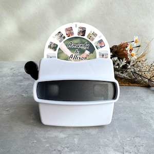 May include: A white View-Master-style viewer with a black eyepiece and a photo reel. The reel displays multiple small photos and the names "Amanda & Allison". The viewer is set against a neutral background with dried flowers.