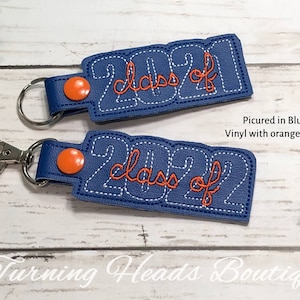 May include: Two blue vinyl keychains with orange snaps. The keychains are decorated with white stitching and the text "Class of 2021" and "Class of 2022" in orange thread.