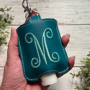 May include: Teal blue leather hand sanitizer holder with a silver clip and a light green embroidered letter "M".