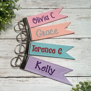 May include: Four personalized keychains with different names, each with a flag-shaped leather tag. The names are Olivia C, Grace, Terence C, and Kelly. The keychains are in pink, peach, blue, and purple.