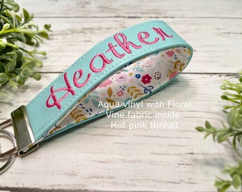 May include: An aqua vinyl wristlet keychain featuring the name Heather embroidered in hot pink thread. The interior is lined with a white fabric displaying a colorful floral and vine pattern. A silver metal clasp secures the loop at one end.