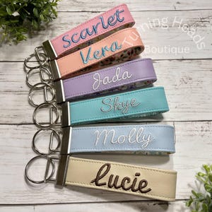 May include: A set of six personalized keychains made of faux leather in pastel colors including pink, peach, lavender, teal, light blue, and cream. Each keychain features a silver metal ring and embroidered names: Scarlett, Vera, Jada, Skye, Molly, and Lucie.