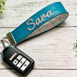 May include: Teal leather key fob wristlet with the name "Sara" embroidered in pink. The wristlet has a floral patterned lining and is attached to a silver key ring with a black car key fob.