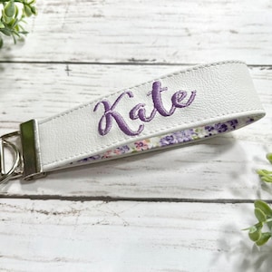 May include: A white faux leather wristlet keychain featuring the name Kate embroidered in purple script thread. The strap includes a silver-toned metal clasp and a circular split ring, with a floral patterned fabric lining in shades of purple and green.