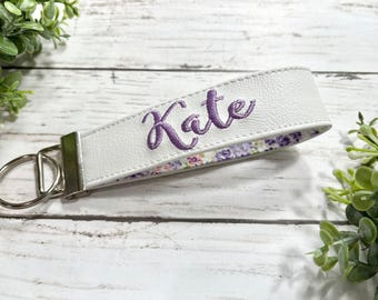 May include: A white faux leather wristlet keychain featuring the name Kate embroidered in purple script thread. The strap includes a silver-toned metal clasp and a circular split ring, with a floral patterned fabric lining in shades of purple and green.