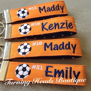 May include: Four orange and black fabric keychains with soccer ball designs and personalized names. The keychains feature the names "Maddy", "Kenzie", "Maddy", and "Emily" with corresponding numbers. The text "Turning Heads Boutique" is printed on the bottom of the keychain.