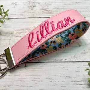 May include: A pink wristlet keychain featuring the name Lillian embroidered in dark pink thread. The strap has a silver metal clasp and a round split ring, with a floral pattern in blue, pink, and yellow visible on the inner side of the loop.