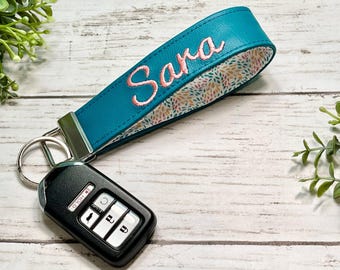 May include: A teal faux leather wristlet keychain featuring the name Sara embroidered in pink thread. The interior is lined with a multi-colored leaf pattern fabric. A silver metal clasp connects the strap to a black car key fob with silver buttons.