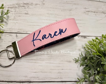 May include: A pink faux leather wristlet keychain featuring the name Karen embroidered in dark blue thread. The keychain includes a silver-toned metal clasp and a circular split ring. The interior lining displays a dark floral pattern.