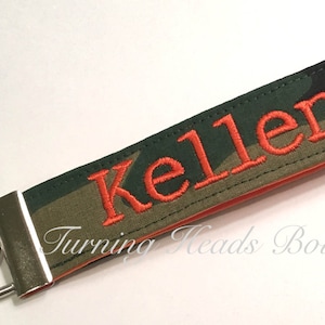 May include: A green and brown camouflage fabric keychain with the name "Kellen" embroidered in orange thread. The keychain has a silver metal ring and a silver metal clasp.