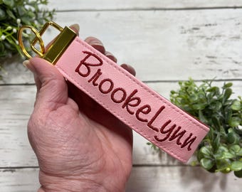 May include: A light pink rectangular keychain strap featuring the name BrookeLynn embroidered in dark red thread. The strap is attached to a gold-toned metal hardware clasp at the top, providing a secure attachment point for keys.