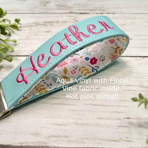 May include: Aqua vinyl wristlet key fob with the name "Heather" embroidered in hot pink thread. The inside features a floral vine fabric. Includes a silver-tone key ring.