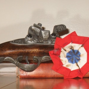 Puede incluir: Una pistola de chispa antigua con una culata de madera oscura y detalles metálicos. Una escarapela roja, blanca y azul se encuentra junto a la pistola, sugiriendo un tema histórico o patriótico. Los objetos descansan sobre una superficie de madera.