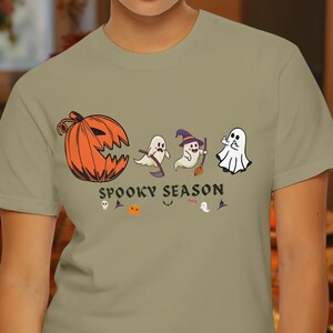 Può includere: T-shirt verde oliva chiaro con grafica a tema Halloween. Il design include una zucca arancione, fantasmi e le parole "Spooky Season" in nero. Sono presenti anche altre icone di Halloween.