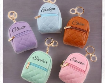 May include: A collection of miniature backpack keychains in various pastel colors, including lavender, light blue, brown, mint green, and pink. Each keychain features a gold zipper and a name printed on the front: Olivia, Evelyn, Chloe, Sophia, and Emma.