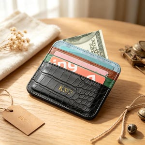 Puede incluir: Un tarjetero negro con textura de cocodrilo y las iniciales KS estampadas en oro junto a un símbolo de corazón. La cartera cuenta con varias ranuras que contienen un billete y varias tarjetas de crédito de colores azul claro, rosa y naranja.